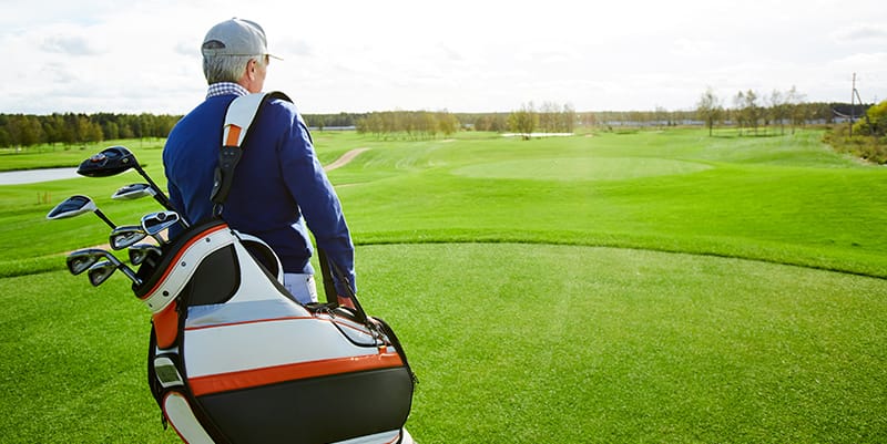 senior man carrying golf clubs while moving along tee box